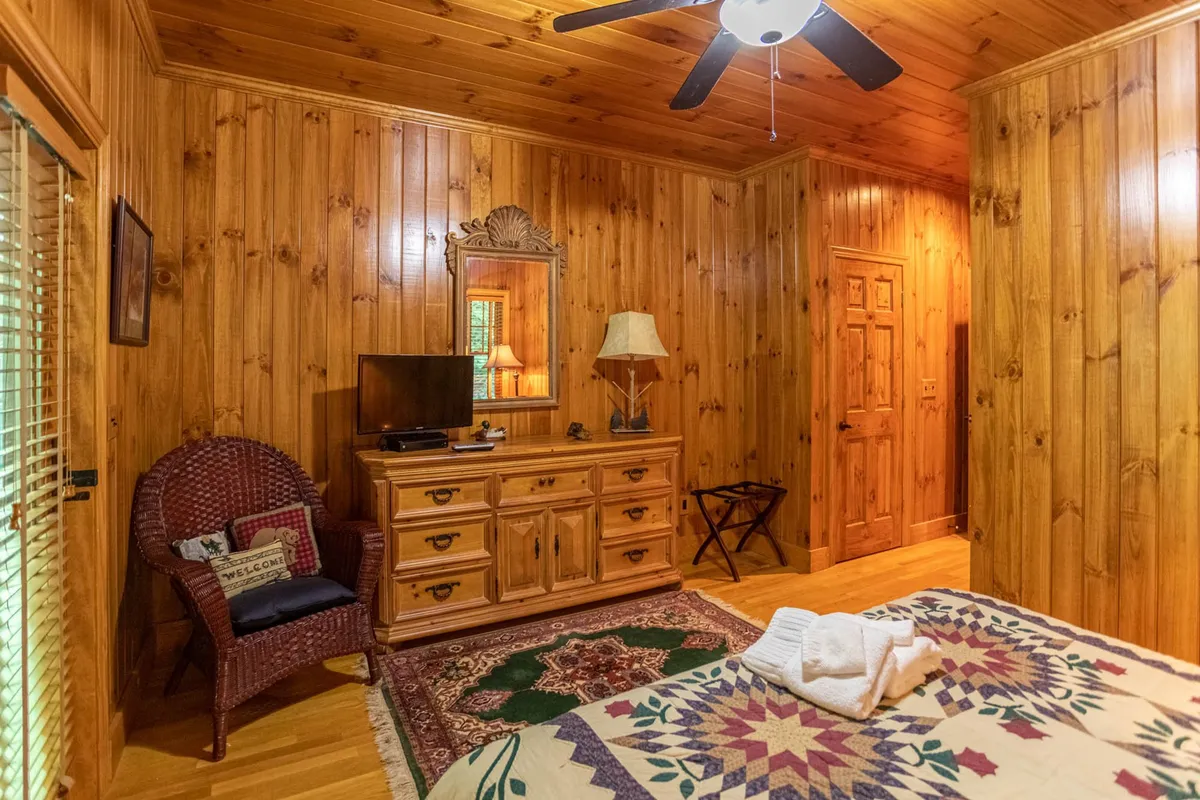 This bedroom scene features a wood-paneled wall, a bed with a cozy quilted bedspread, a wooden dresser, and a woven chair beneath a warm lamp. The overall mood is restful and inviting, ideal for privacy and unwinding.