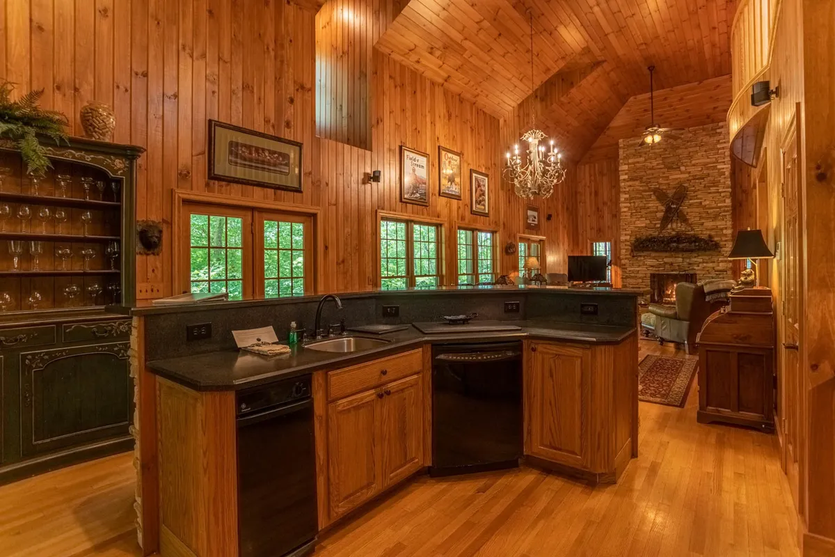 The image spotlights a kitchen with a prominent stone island, adjacent to both a stone fireplace and wooden cabinetry. Large windows brighten the space, and a chandelier adds elegance. The island is central and generously sized, making it ideal for prepping meals, socializing, or enjoying coffee in the morning light.