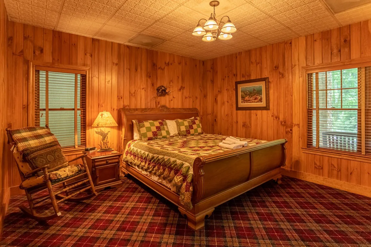 A bedroom with a bed, rocking chair, lamp, and plaid carpet. The window allows for views of the outdoors, inviting guests to enjoy a book or the sunset through the trees, which ties back to explorations and moments of quiet reflection.