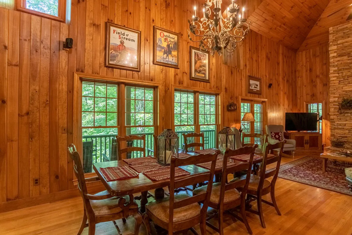 This image centers on a warmly lit dining area with a long table under a chandelier, surrounded by wood paneling, large windows, and a stone fireplace. The setting is intimate yet spacious, inviting both family meals and festive celebrations.