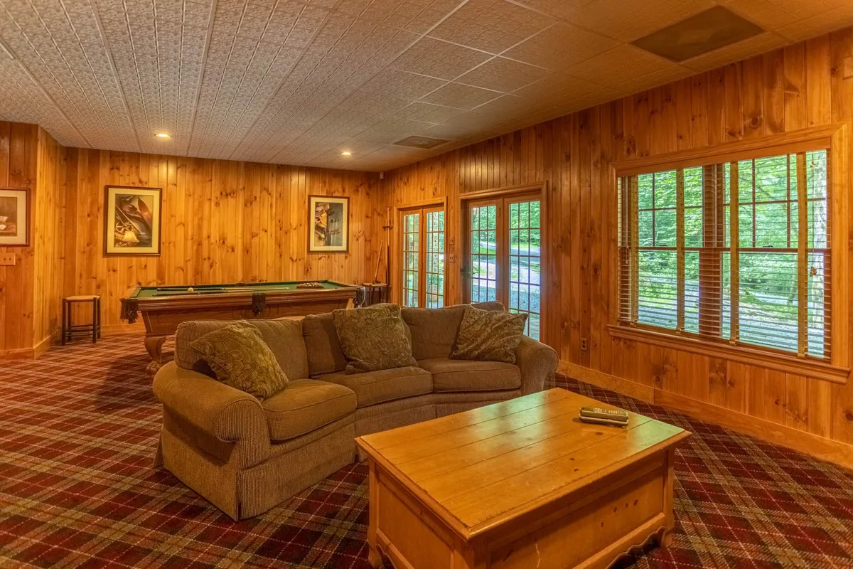 An inviting lower-level family room with a comfortable couch, pool table, wooden coffee table, and large windows bringing in natural light. The space feels perfect for game nights or friendly competition among family and friends, with a relaxed and informal vibe.