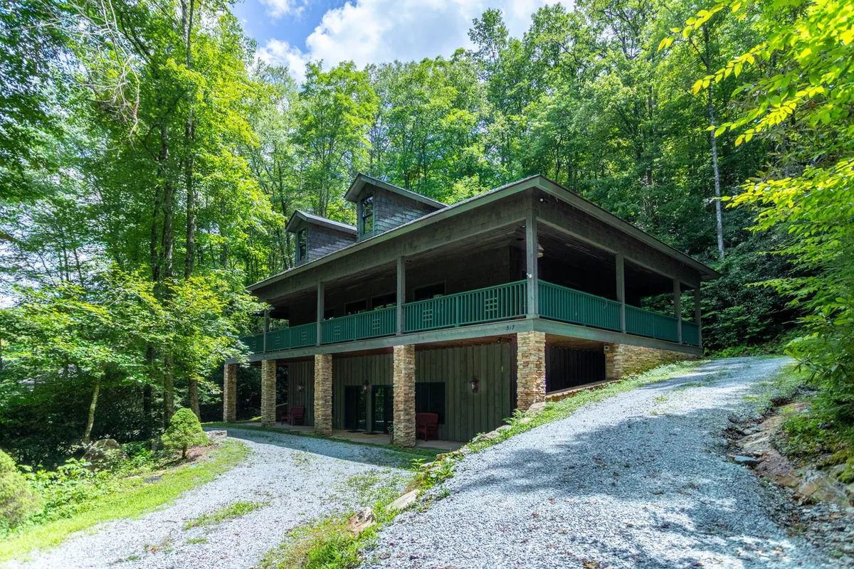 The image showcases the cabin's striking exterior, featuring a welcoming, expansive porch perfect for relaxing, a sturdy stone foundation, and lush woodland surroundings that create a tranquil and private atmosphere.