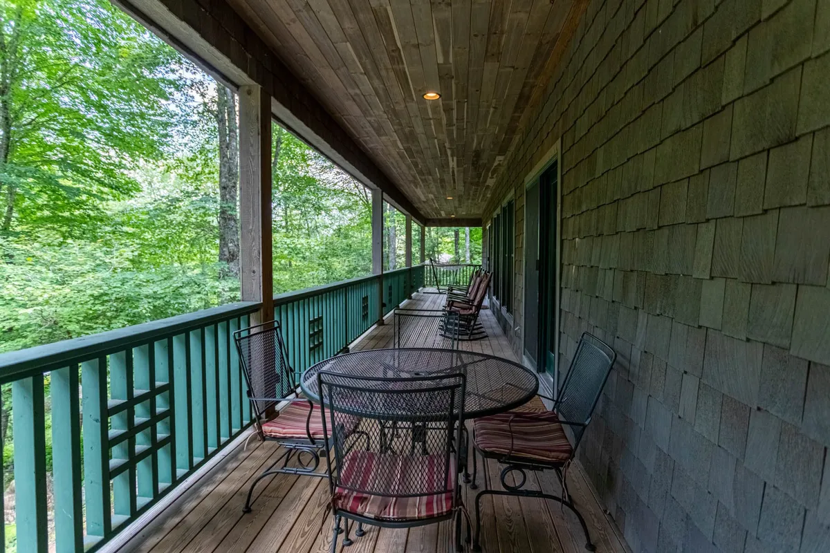 A wide, welcoming porch with a circular table surrounded by chairs, set against a backdrop of lush greenery and woodland. The setting is tranquil, perfect for relaxing with a book, sipping morning coffee, or gathering with friends at the edge of nature.