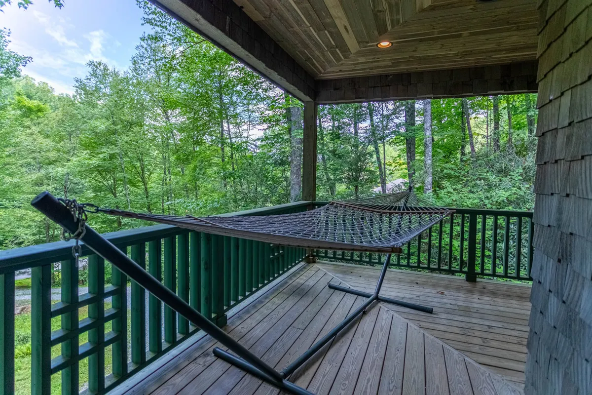 A peaceful exterior shot featuring a hammock on a wooden deck, surrounded by lush greenery. The area is ideal for relaxation, reading, or stargazing, with hints of morning or sunset light filtering through the trees.
