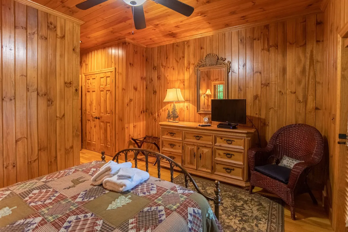 A cozy bedroom with wood-paneled walls, a patterned quilt on the bed, dresser with a mirror, lamp, television, and a wicker chair. The space exudes comfort and provides an inviting retreat for rest after a day of fun.