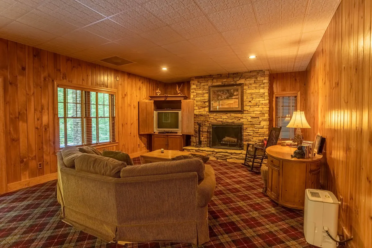 A warmly lit den featuring a fireplace, comfortable seating, wood-paneled walls, and windows revealing woodland scenery outside. The space is designed for intimate gatherings or quiet relaxation, echoing the serenity described in the section.