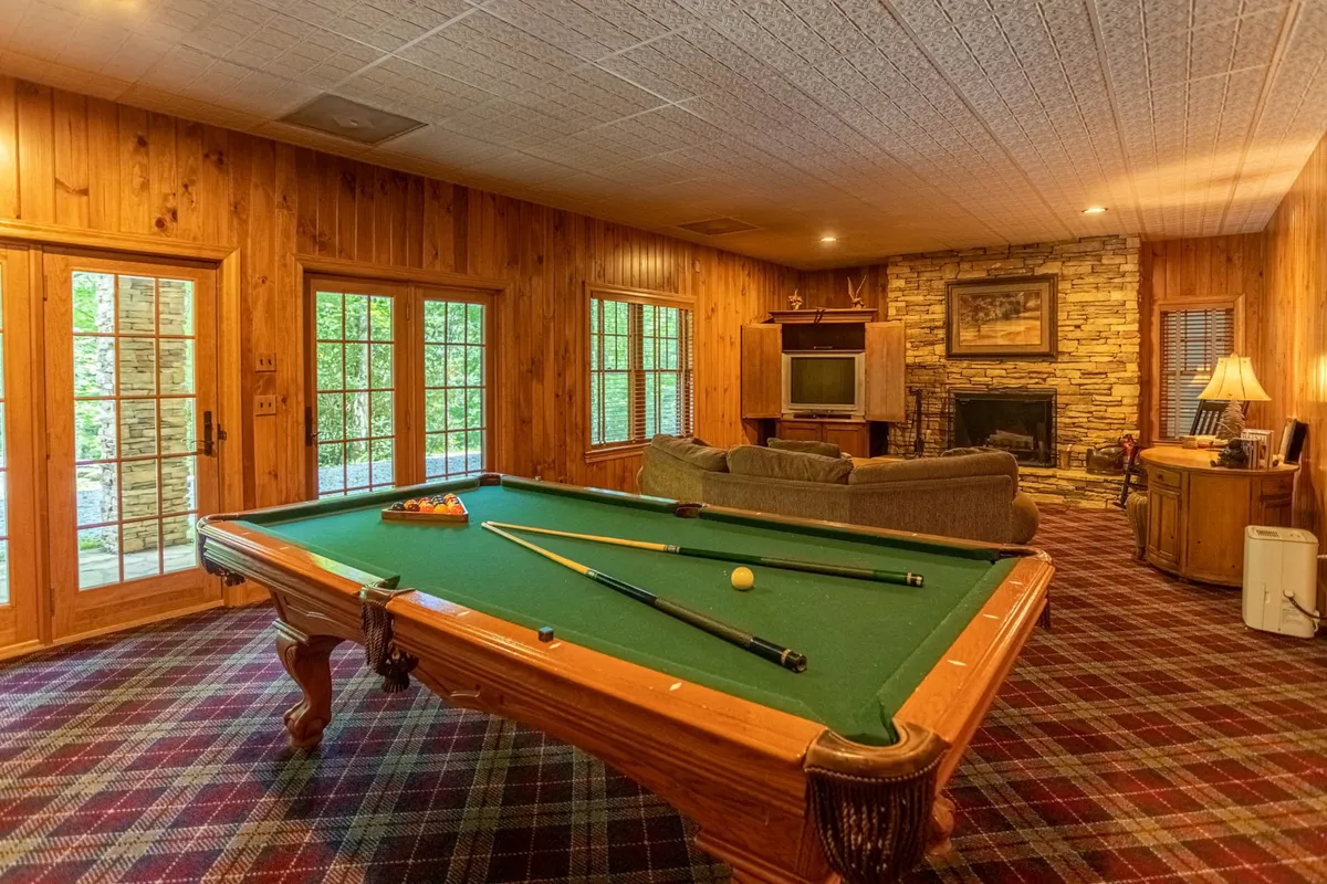 This image features a family-friendly game room with a pool table at the center, a comfortable couch, a stone fireplace, and windows bringing in natural light. The space clearly invites group activities, relaxation, and lively gatherings by the fire.