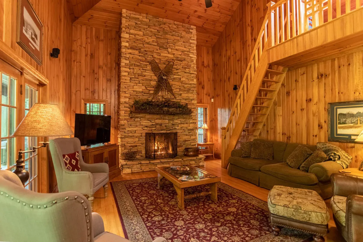This image showcases a warmly inviting great room featuring a striking stone fireplace, a wooden staircase, and plush, comfortable seating arranged on a decorative rug. The ambiance is cozy and perfect for socializing or relaxing after a day exploring the area.