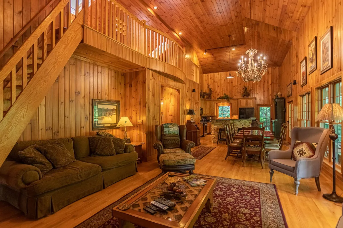 This image captures a spacious, open-concept living and dining area. The scene features a sweeping wooden staircase, a central chandelier, a comfortable sofa seating area, and an inviting dining table. Natural light pours in through large windows, and the space feels relaxed and welcoming—ideal for family gatherings and cozy evenings.