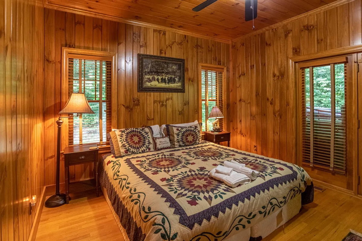 A warmly lit bedroom with a large bed topped with a cozy quilt, flanked by bedside lamps and surrounded by inviting wood paneling. Large windows let in gentle daylight and provide views of the surrounding greenery, creating a serene atmosphere for rest and relaxation.