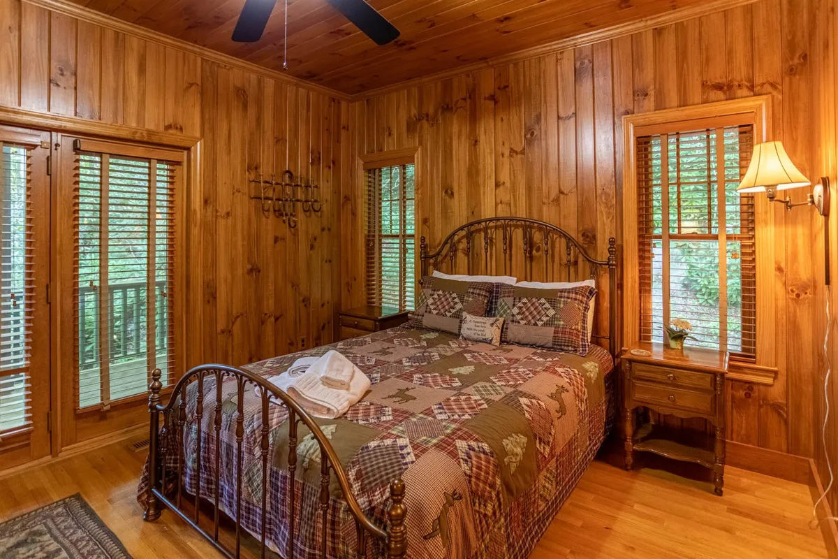 A spacious bedroom featuring a bed with a quilted cover, natural wood walls, large windows that invite in the forest scenery, and a nightstand with a lamp. The setting is cozy yet generously sized, offering both comfort and a sense of seclusion.